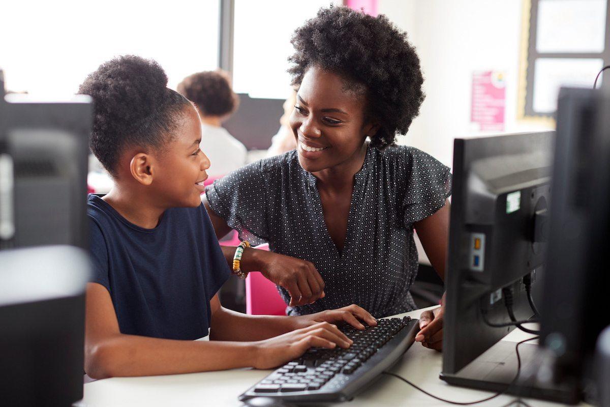 A Black educator smiles at a Black middle school student as they work 1-on-1 together in their school's computer lab.