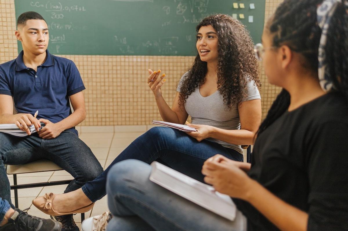 Three high school students sitting in a classroom, having a discussion. A student with long curly hair has a book in her lap and is sharing an idea while her classmates listen closely.