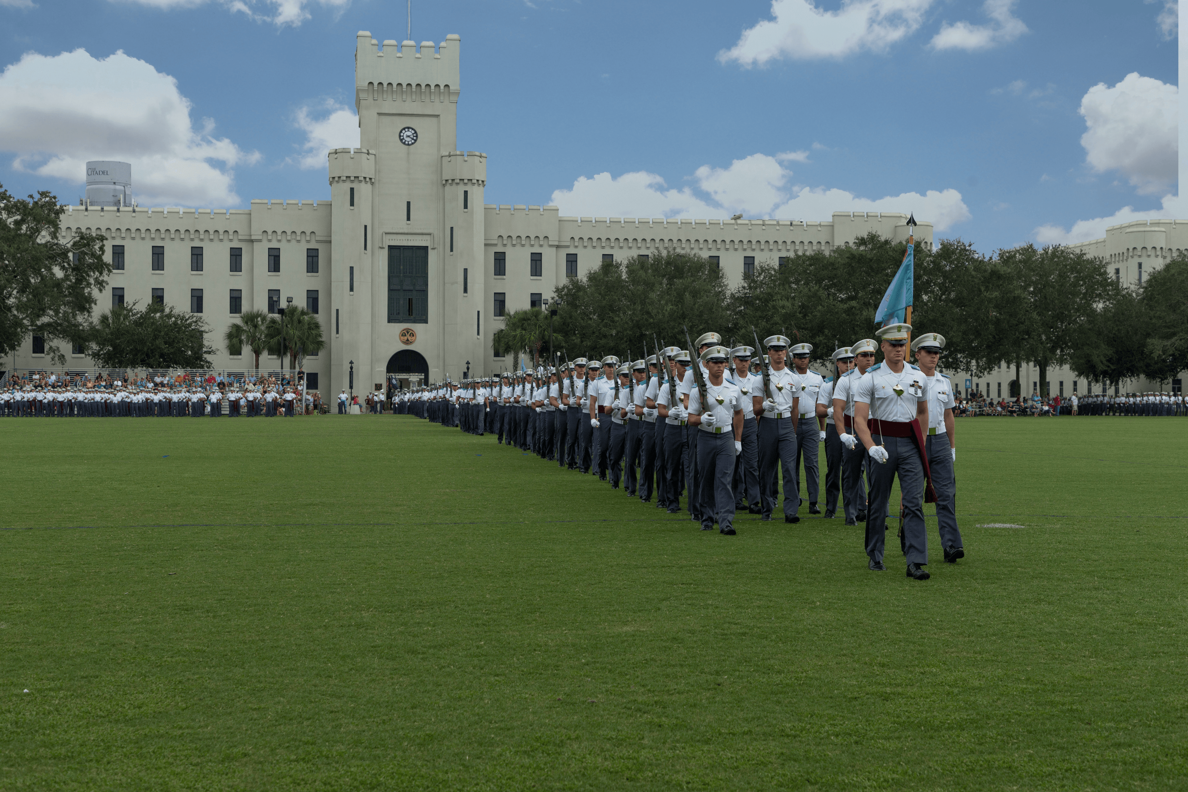 Cadets wearing white uniform marching in front of school building.