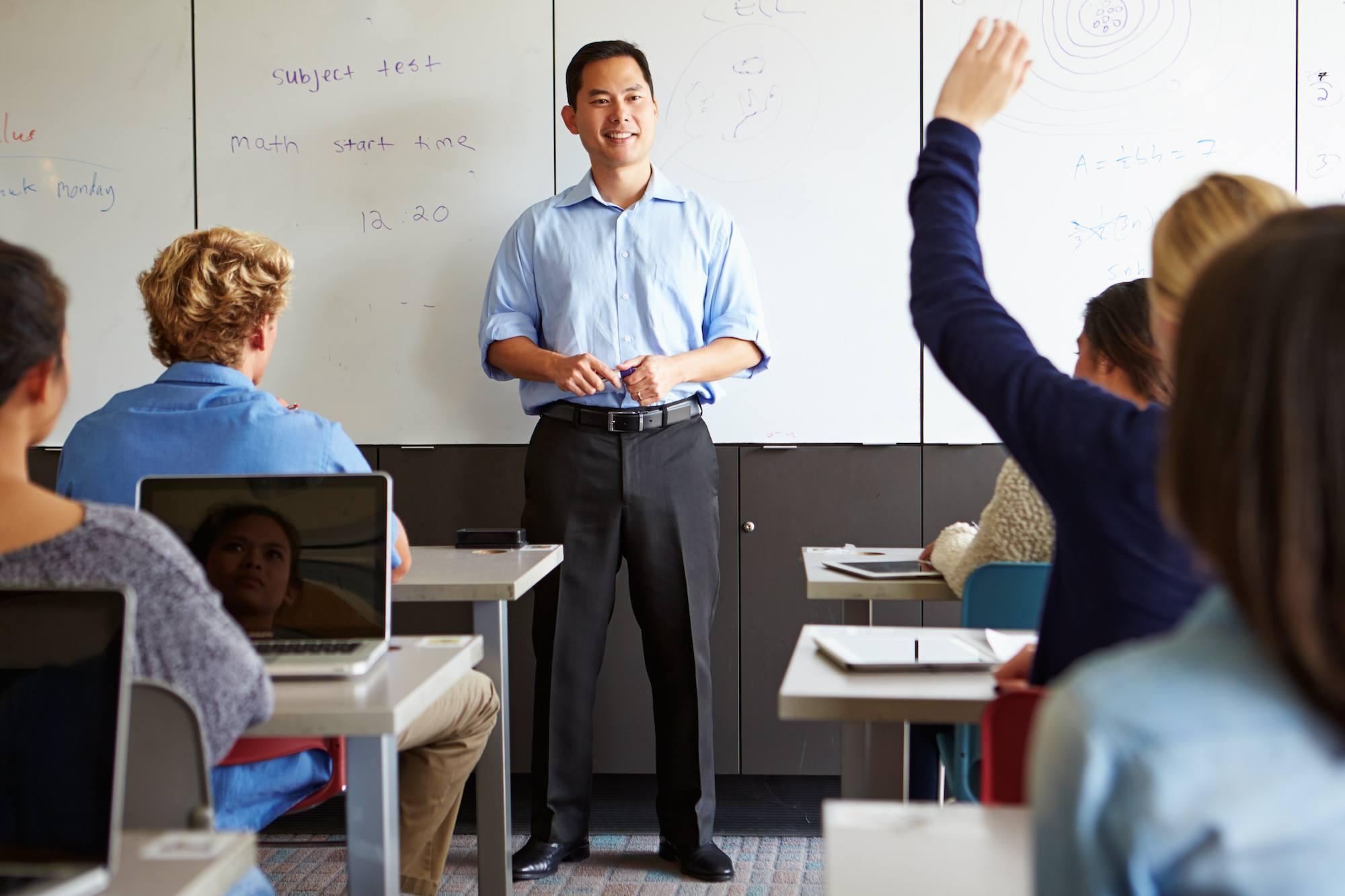 A high school math teacher leads a lesson at the whiteboard. He smiles a student raising their hand.